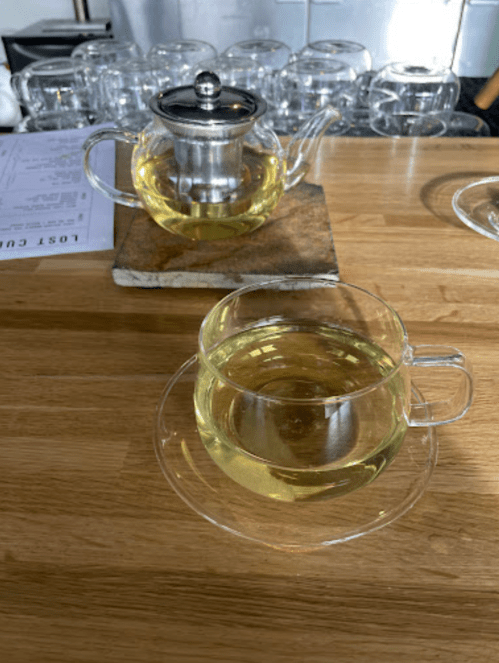 A clear glass teacup filled with light yellow tea sits on a wooden table next to a teapot and a coaster.